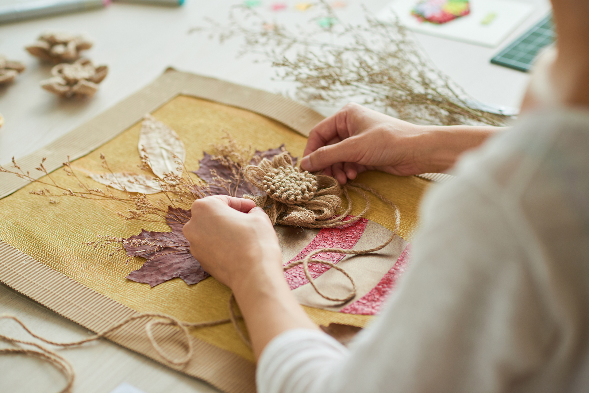 Over the shoulder image of a person constructing a collage with various elements such as yarn, dried flowers, and seed pods. 