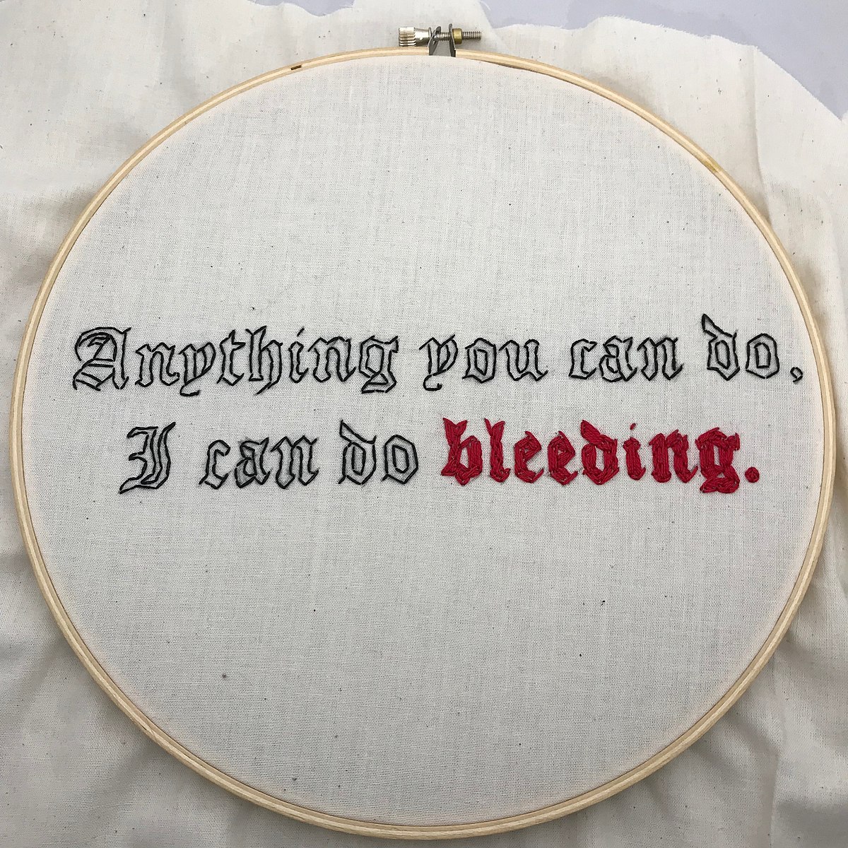 A cross stitch that reads "Anything you can do, I can do bleeding", with the word bleeding in red thread in white linen cloth .