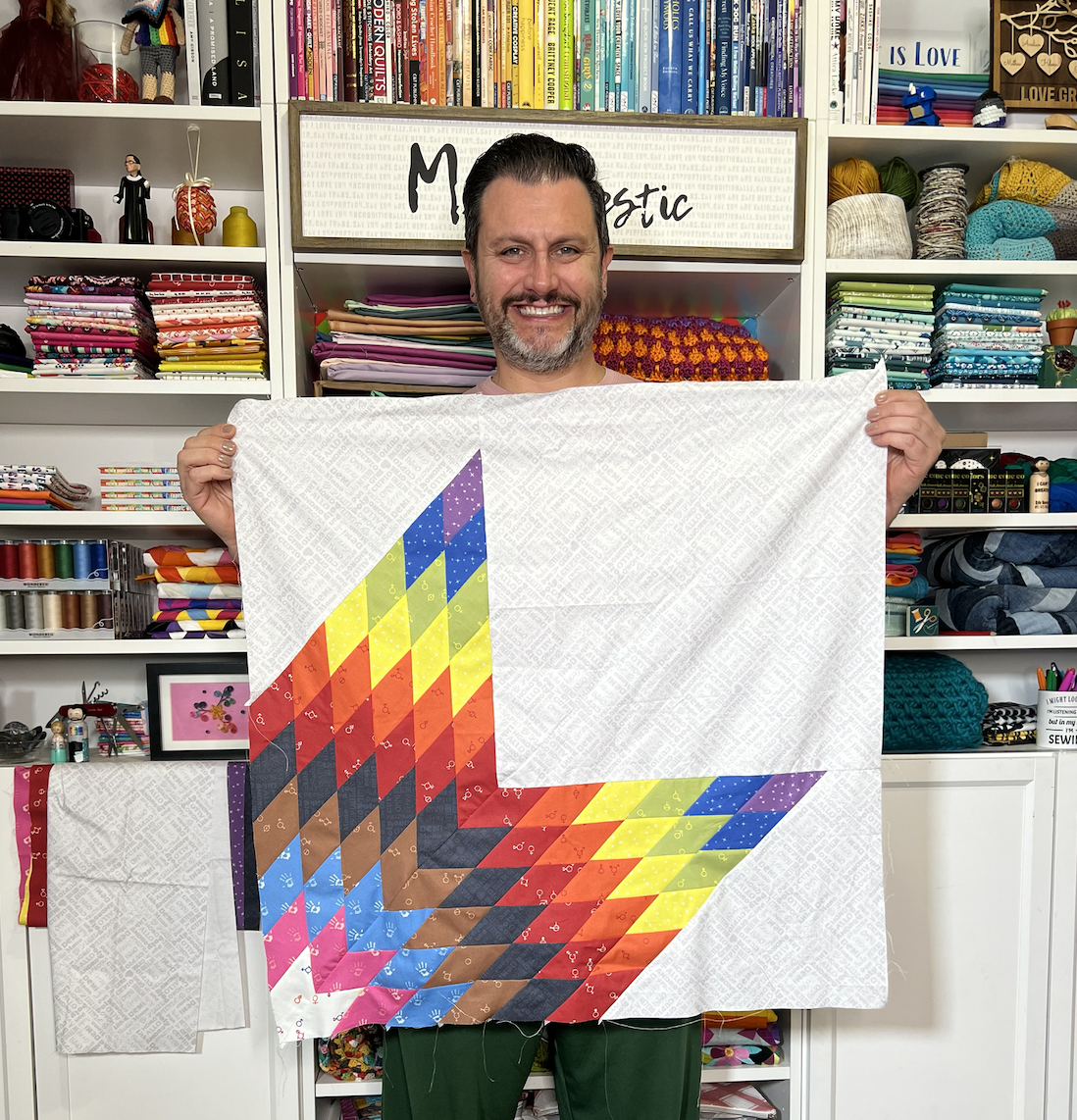 Person with goatee and dark hair holding up a quilt with 2 points crafted in the LGBTQ+ rainbow flag.