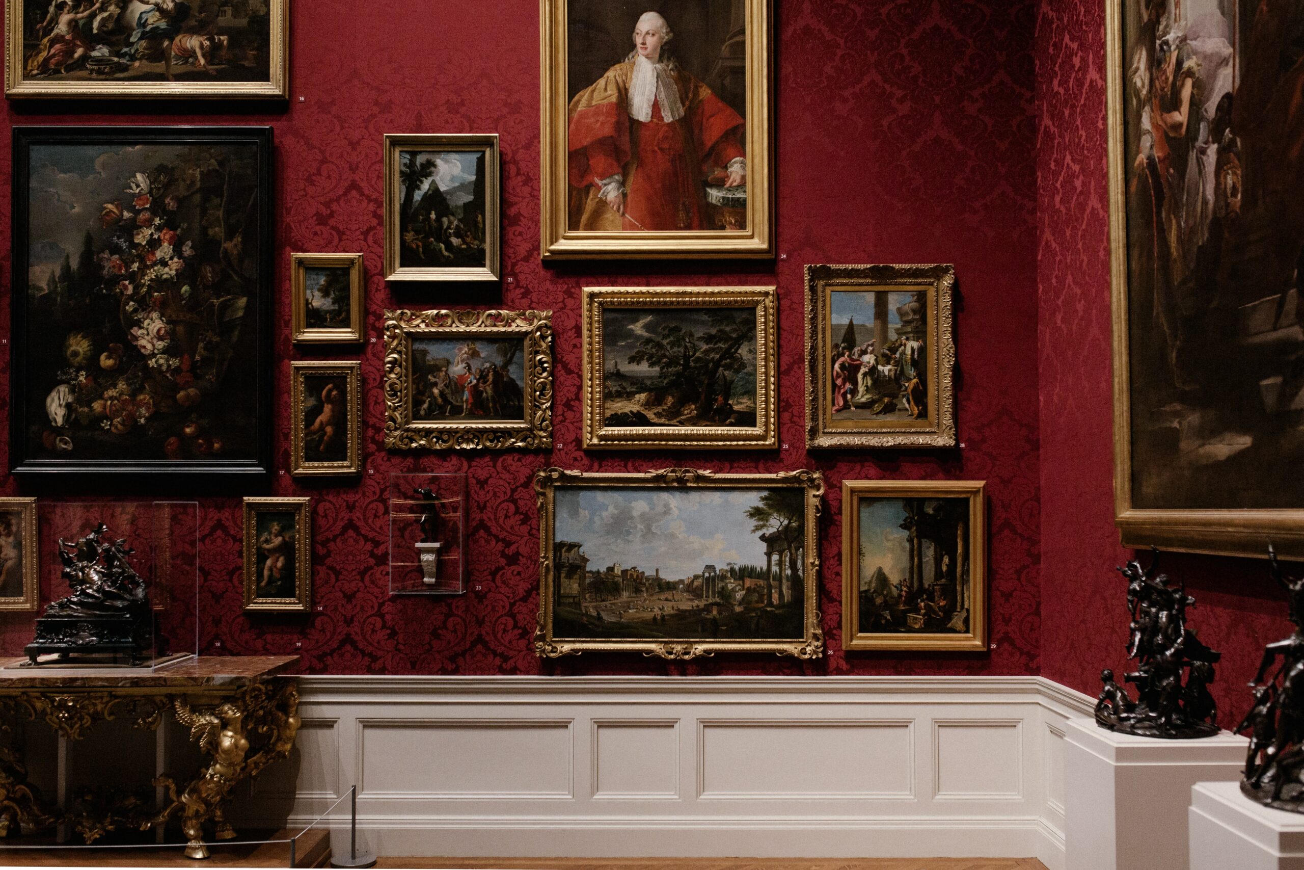 A museum wall with paintings above a table with sculptures.