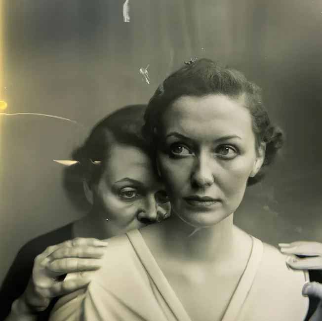 Sepia tone photograph of two white women with brown hair. Woman in front looking off camera to her right, woman behind has her hands on the shoulders of the woman in front while seemingly resting her mouth on her back while her eyes gaze calmly down but in front of the woman in front.