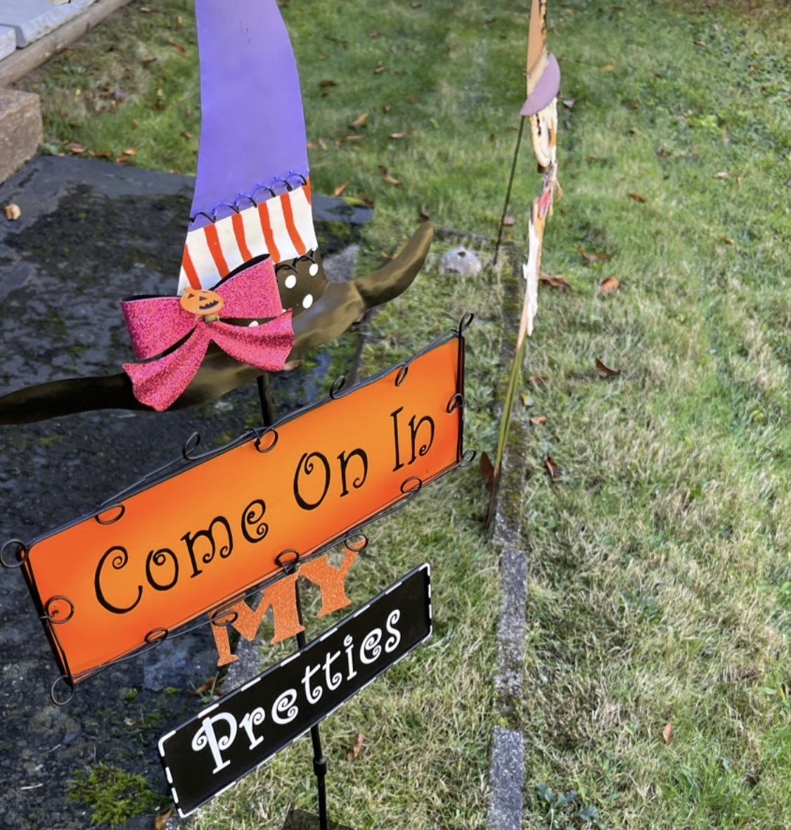 A Halloween sign stuck in a yard with a purple witch hat above a sign saying, "Come on in my pretties"