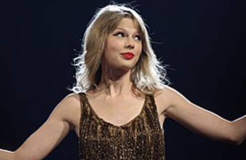 A photo of Taylor Swift wearing a shiny gold tank top in concert, photographer from the waist up with a blank, black background.