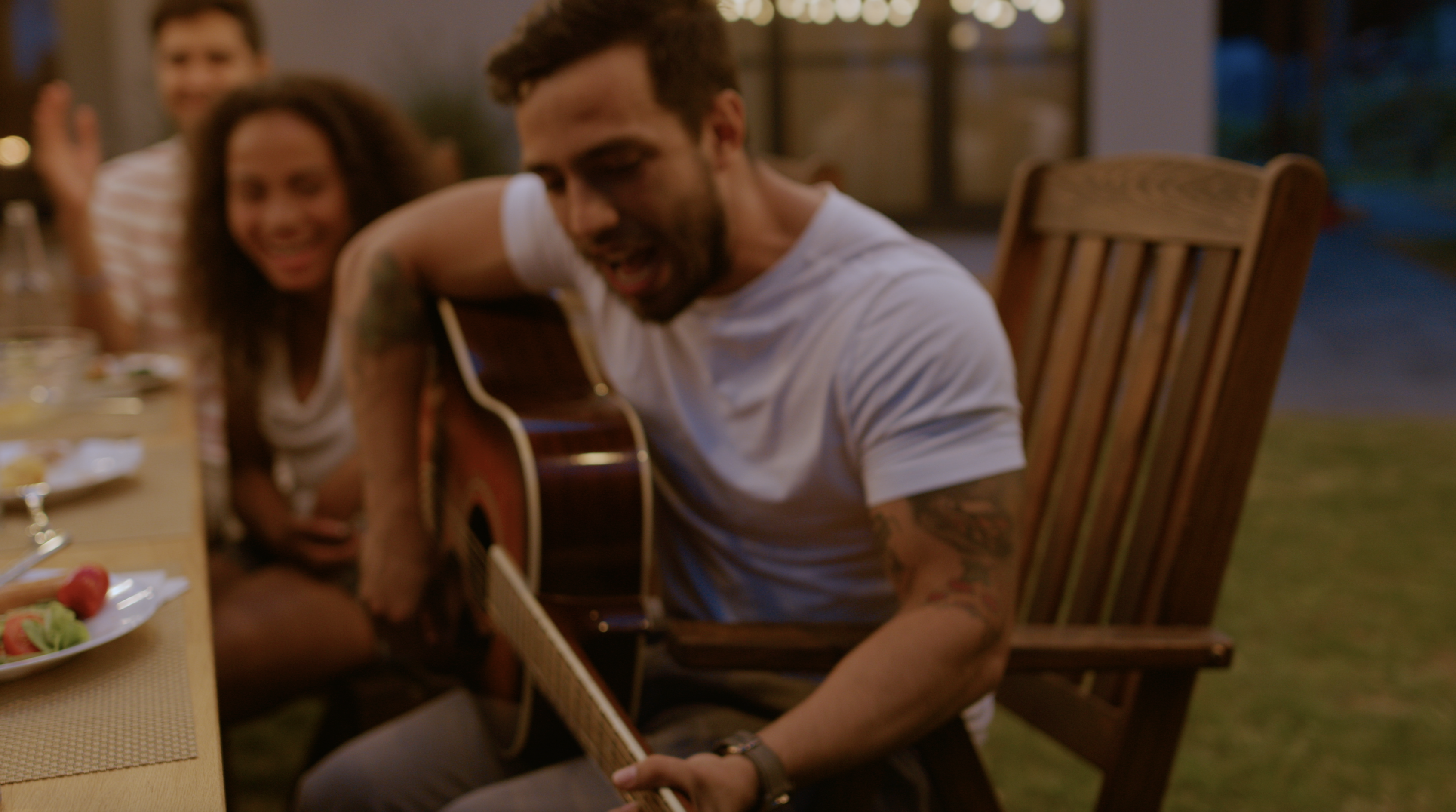 A young man with dark hair plays the guitar while seated at a dinner table. Family members smile at him in the background.