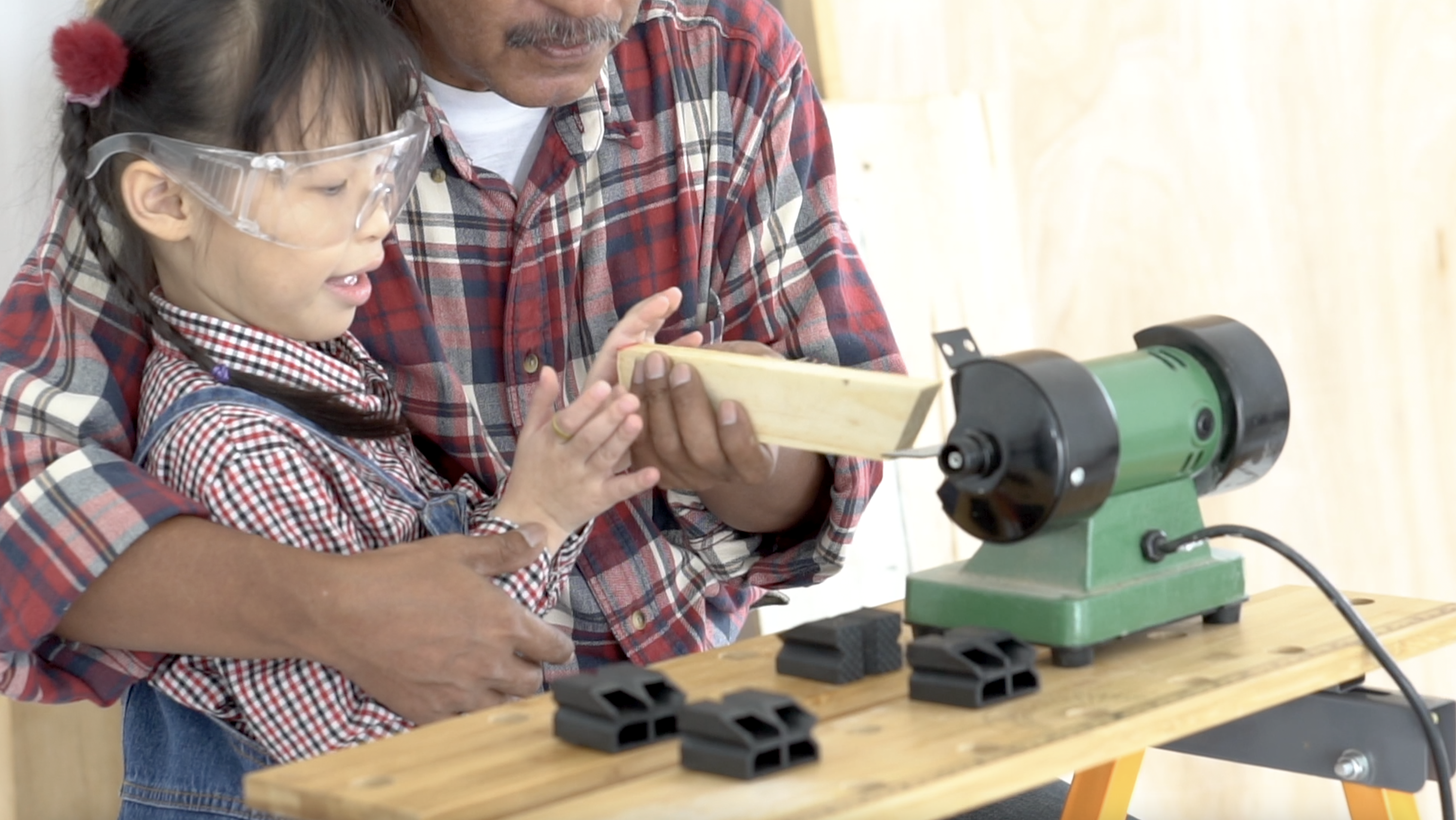 A young Asian girl wears safety glasses and sits in the lap of her father. They look at a piece of wood while woodworking equipment is nearby on a table.