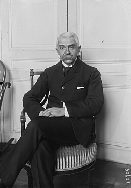 A black and white photo from 1921 of a light-skinned man with white hair seated on a wooden chair against a white wall. His legs are crossed, his hands on his knee, and he stares without expression at the camera.