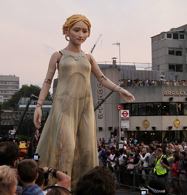 A photograph of an over-sized human puppet of light skinned, blonde puppet in a white gown, parading during the Cultural Olympiad Carnval in the London 2012 Games.