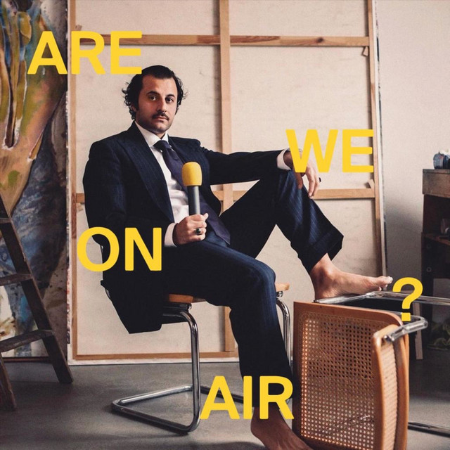 A man in a suit sits barefoot on a modern chair in an art studio. He holds a yellow microphone, looking at the camera with a thoughtful expression. The words "ARE WE ON AIR?" are overlaid in bold yellow text. The background features a canvas and artistic materials.